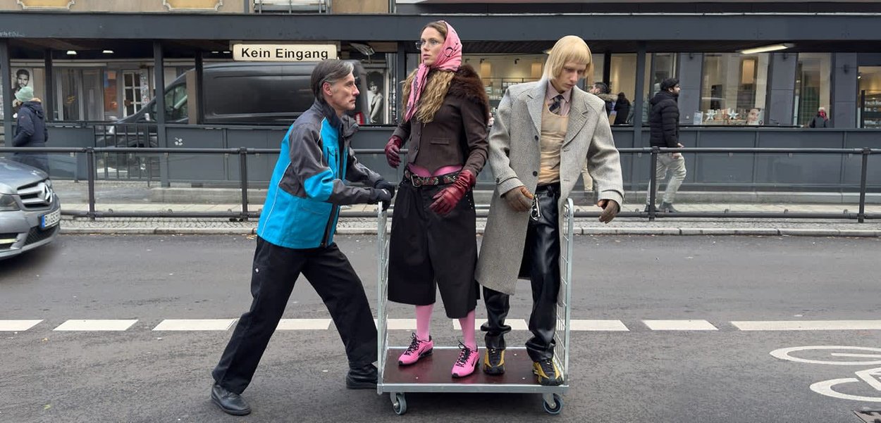 Två skyltdockor på en vagn: en i en brun kappa och rosa halsduk, den andra i en ljusgrå kostym. En person skjuter vagnen på en gata.