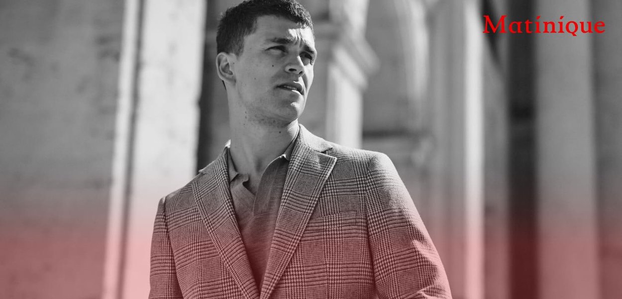 Blazer grigio a quadri con revers appuntiti, abbinato a una camicia chiara. Lo sfondo presenta elementi architettonici. Sovrapposizione di testo in rosso.