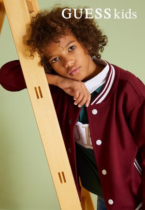 Un bambino con i capelli ricci indossa una giacca varsity color bordeaux e si appoggia a una scala di legno su uno sfondo verde chiaro.
