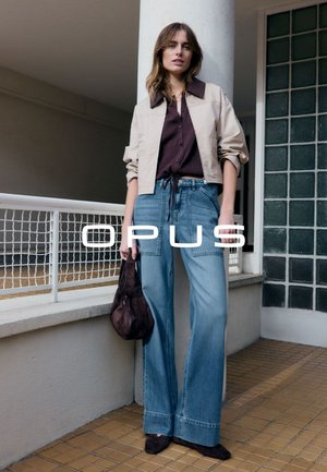 Femme portant une veste beige courte, une chemise marron foncé, un jean bleu à jambes larges, tenant un sac marron foncé, debout à l'intérieur près d'une rambarde.