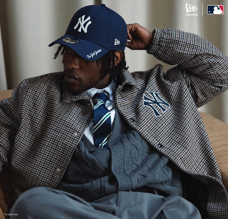 Uomo che indossa un cappello blu dei New York Yankees, giacca a quadri con logo Yankees, cardigan grigio, cravatta a righe, reclinato con la mano dietro la testa.