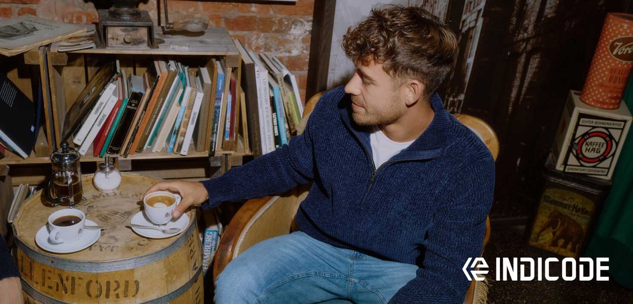 Strickpullover in Marineblau, helles Hemd und blaue Jeans, getragen von einem sitzenden Mann. Zwei Kaffeetassen auf einem hölzernen Fass-Tisch, im Hintergrund Bücher.