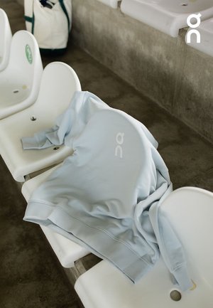 Light blue sweatshirt laid on a white stadium seat, featuring a subtle logo in the center, with long sleeves and a soft texture.
