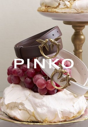 Due cinture, una marrone e una bianca, ciascuna con una fibbia circolare dorata decorata con accenti a forma di uccello, esposte su una torta guarnita con uva.