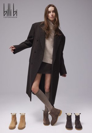 Manteau marron en laine croisé, pull en maille beige, jupe sombre, chaussettes montantes ; bottes beiges et sombres avec doublure en fourrure et semelles jaunes.