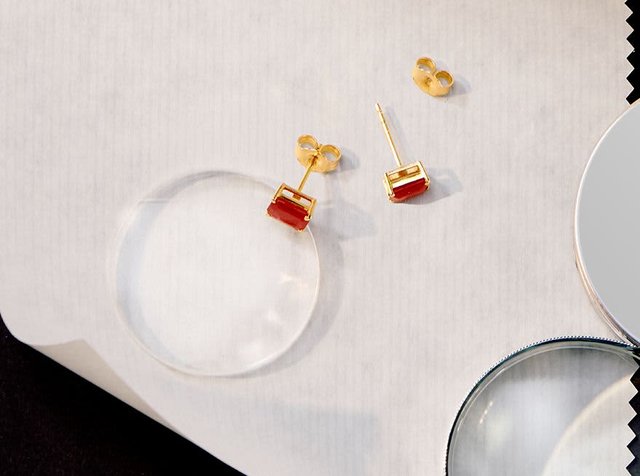 Χρυσά σκουλαρίκια με ορθογώνιες κόκκινες πέτρες τοποθετημένα σε λευκή επιφάνεια δίπλα σε δύο μεγεθυντικούς φακούς.