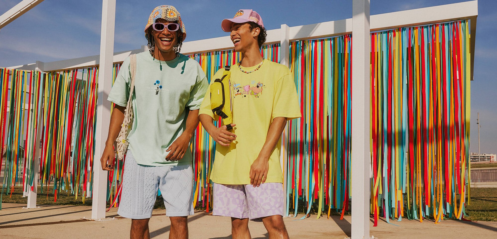 Twee mannen staan naast elkaar, gekleed in pastelkleurige overhemden en gestreepte shorts, met kleurrijke lintdecoraties op de achtergrond. Accenten omvatten hoeden en een crossbody tas.