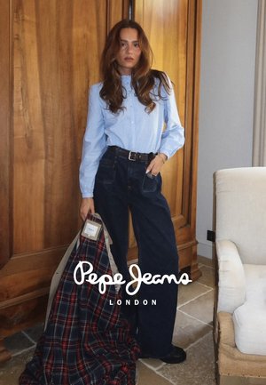 Model draagt een lichtblauw button-up shirt en donkerblauwe wijde jeans. Ze houdt een geruite stof vast, tegen een houten wand.