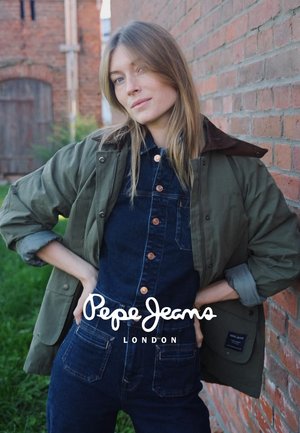 Parka verte avec col marron superposée sur des salopettes en denim foncé avec des boutons et des poches orange. Mur en briques en arrière-plan.