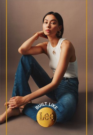 Hvid ribbet tanktop kombineret med blå denim jeans. Modellen sidder i en afslappet pose, og viser et gyldent rundt vedhæng og store øreringe.