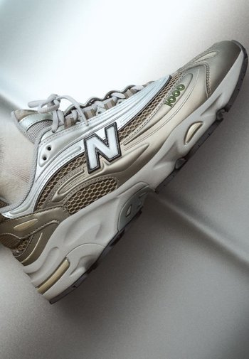Beige und silberner Sneaker aus Mesh- und Leder-Materialien. Besitzt ein auffälliges schwarzes "N"-Logo, strukturierte Akzente und eine gepolsterte Sohle.