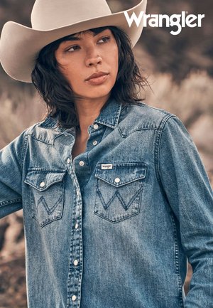 Light blue denim shirt with two chest pockets and zigzag stitching, featuring a button-up design and a brand label. Model wears a wide-brimmed hat.