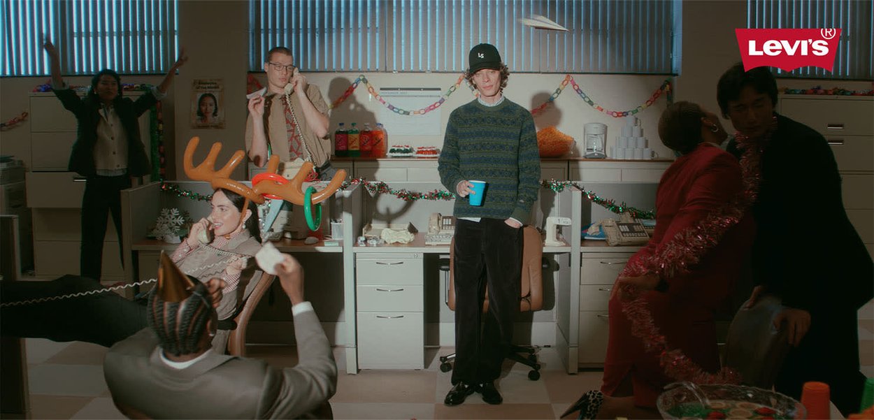 Office scene with festive decorations. People wearing varied clothing, including reindeer antlers, engaged in playful activities amid a cluttered workspace.