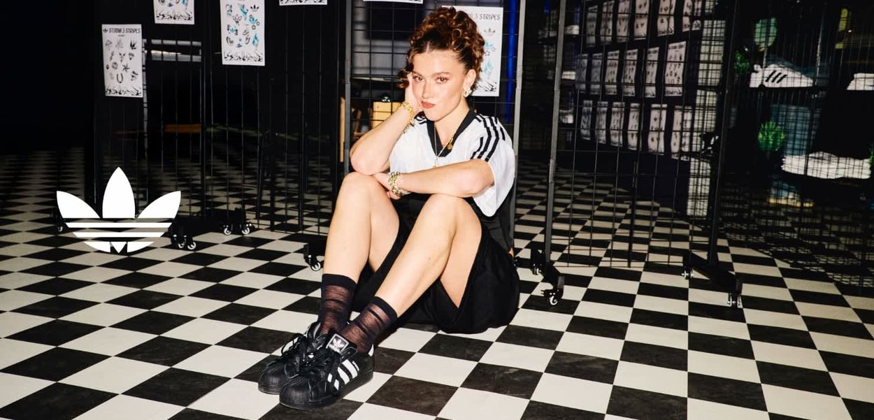 Woman in Adidas sportswear sitting on black-and-white checkered floor inside a store with tattoo design displays on metal grids.