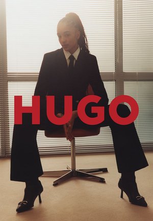 Abito nero con camicia bianca, seduto su una sedia arancione. Scarpe con tacco alto con un accento decorativo. Testo "HUGO" in rosso audace.