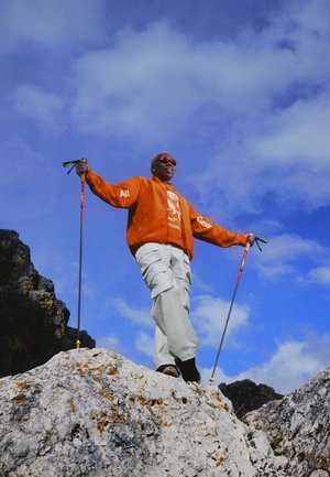 Persona con giacca arancione che tiene bastoncini da trekking in piedi su terreno roccioso sotto un cielo azzurro con nuvole sparse.
