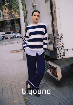 Femme appuyée contre un camion sur un trottoir en ville, portant un pull rayé bleu marine et blanc avec un pantalon bleu marine et des chaussures noires.