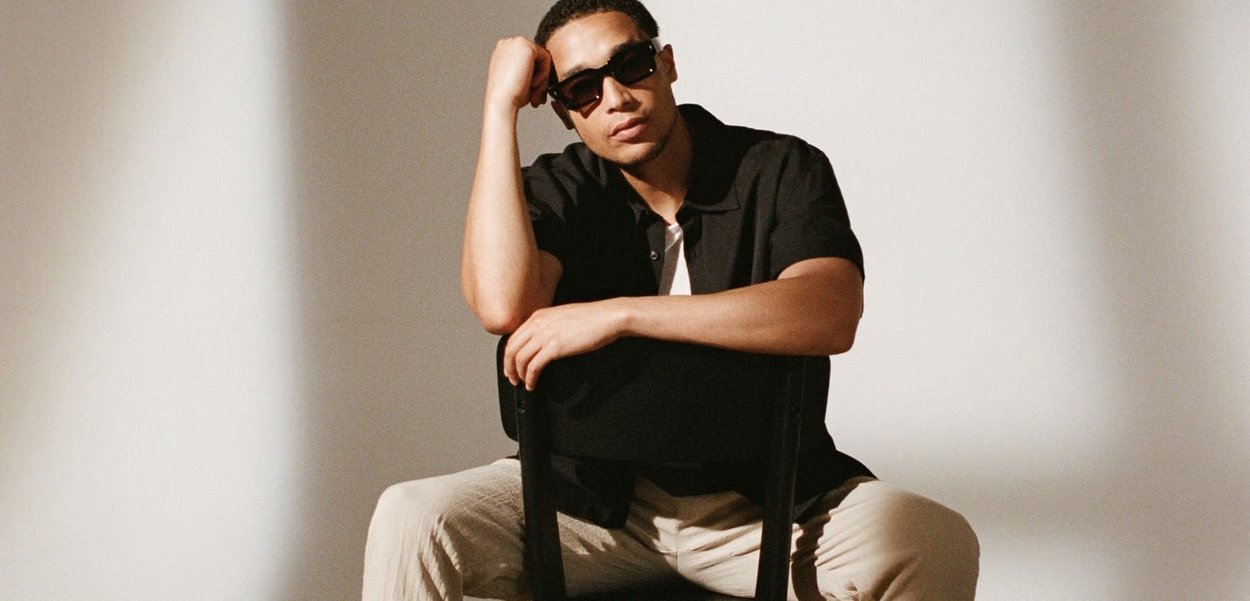 Young man in sunglasses sits backwards on a chair, resting his arm on the chair back in a neutral, light-filled space.