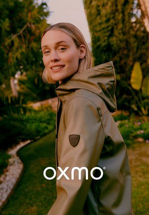 Ung kvinde iført en olivengrøn regnjakke med hætte smiler udendørs i en have med træer og planter ved solnedgang.