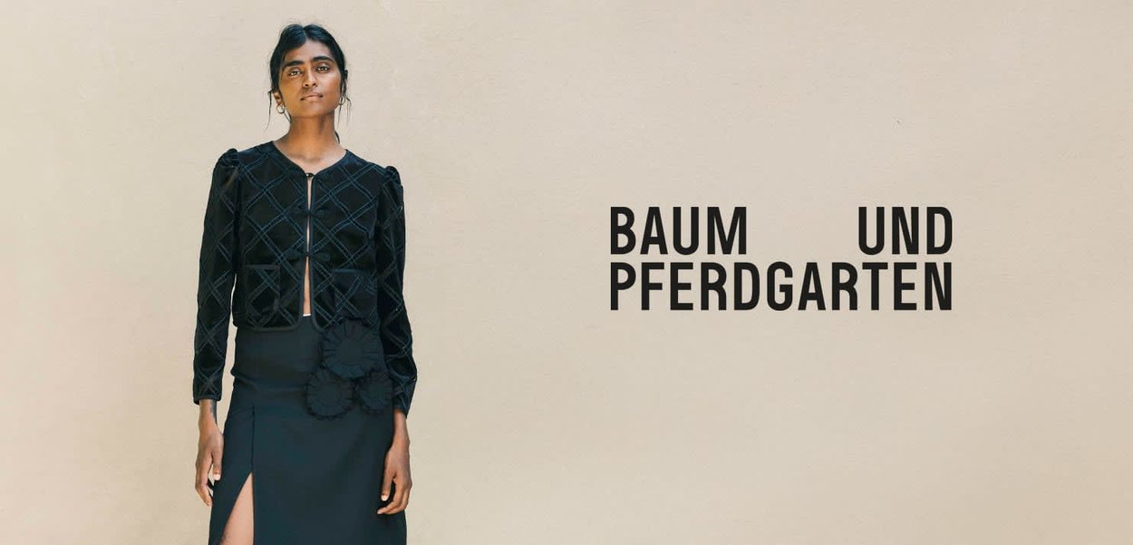 Kobieta w czarnej, teksturowanej kurtce i spódnicy z detalem kwiatowym stoi obok napisu "BAUM UND PFERDGARTEN" na beżowym tle.