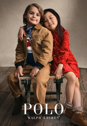 Beige Cordjacke, marineblauer gemusterter Pullover, rotes Kleid mit Pünktchen. Kinder sitzen auf hölzernem Hocker, braune Stiefel, strukturierter grauer Hintergrund.