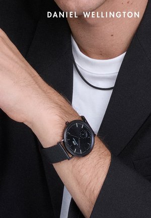 Czarny zegarek z okrągłą tarczą, siateczkowym paskiem oraz białymi i niebieskimi akcentami. Noszony na nadgarstku, zestawiony z ciemnym żakietem i białą koszulką.