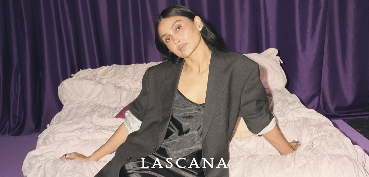 Czarny żakiet na ciemnej sukience z satyny, siedząca na pomarszczonej jasnej pościeli, w tle fioletowe zasłony. Wyraźne logo na dole.