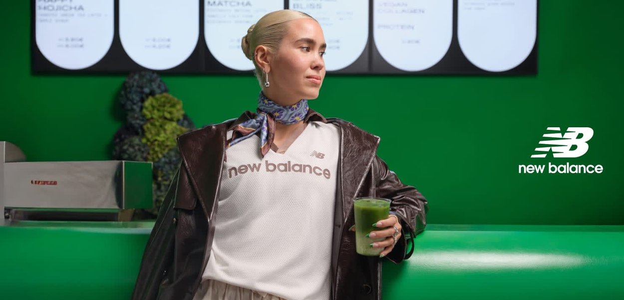 T-shirt en mesh blanc avec le texte « new balance », veste en cuir marron foncé, écharpe colorée, tenant une boisson verte, sur un fond vert vif.