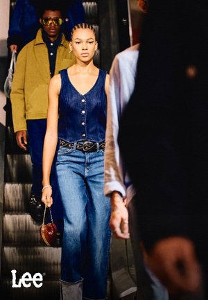 Denim jurk met knopen, gecombineerd met wijde blauwe jeans. Een zwarte riem met een decoratieve gesp en een kleine handtas in de vorm van een hart.