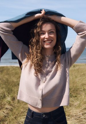 Junge Frau mit lockigem Haar lächelt und hält eine Jacke über ihren Kopf in der Nähe eines grasbewachsenen Strandes und des Ozeans unter klarem Himmel.