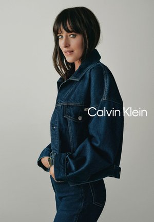 Mujer de cabello oscuro hasta los hombros, vestida con una chaqueta de mezclilla azul oscuro y pantalones vaqueros, de perfil frente a un fondo liso.