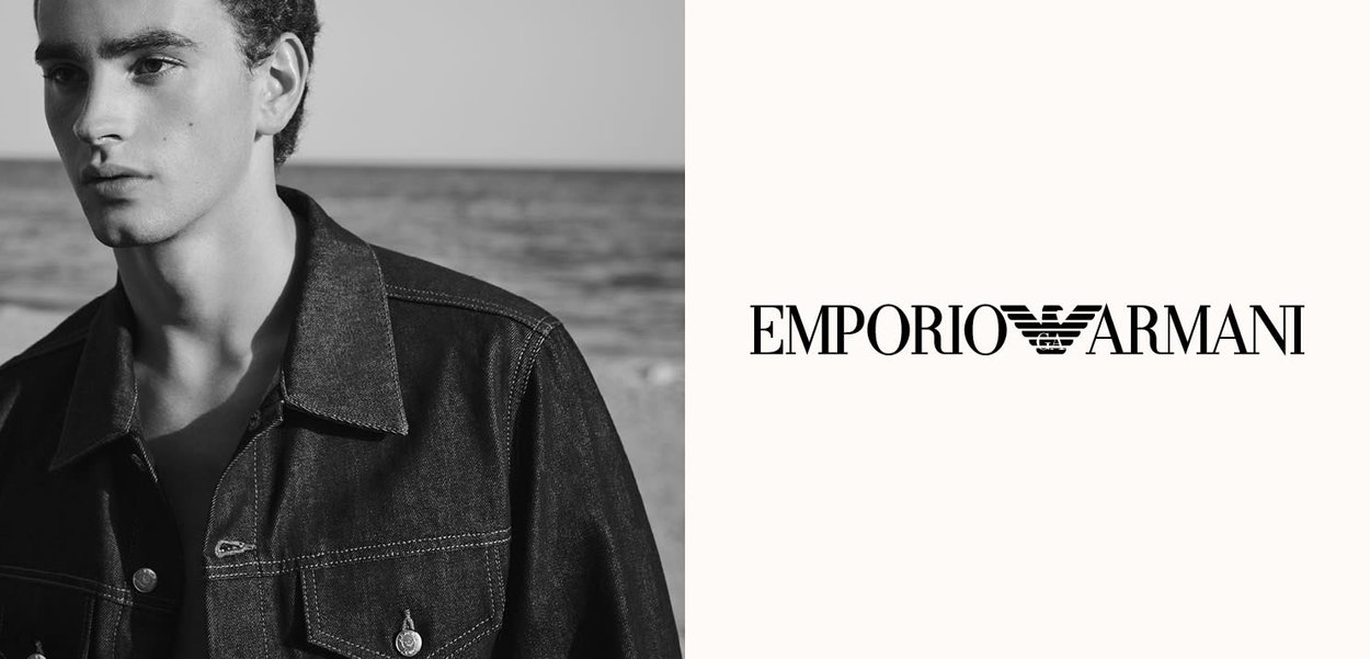 Joven con chaqueta de mezclilla oscura junto al mar, al lado del logo negro de Emporio Armani sobre fondo blanco.