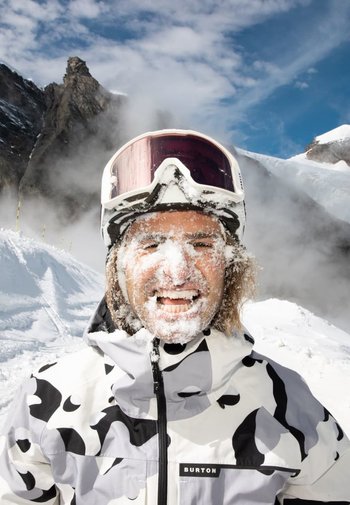 Die Skijacke hat ein Schwarz-Weiß-Muster in Kuhflecken und eine glatte Textur. Die Person hat Schnee im Gesicht, trägt Skibrillen und grinst.