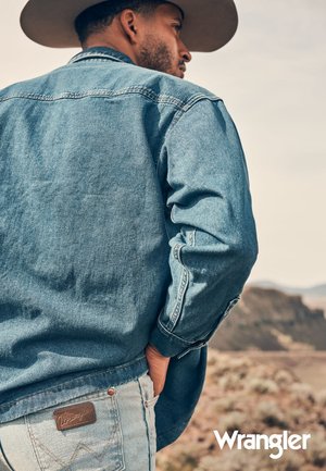 Lys blå denimjakke med knappåpning og to brystlommer, kombinert med falmede blå jeans med et brunt Wrangler-skinnlapp.