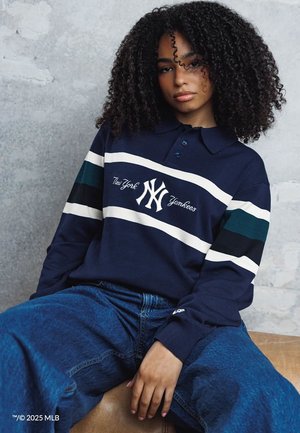 Giovane donna con capelli ricci che indossa una maglia a maniche lunghe blu scuro dei New York Yankees e jeans blu, seduta contro un muro grigio dalla superficie texturizzata.
