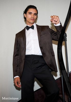 Brauner Wildleder-Blazer über einem weißen Hemd, schwarzer Fliege und schwarzen Hosen. Model posiert auf einer Treppe mit einer strukturierten Wand im Hintergrund.