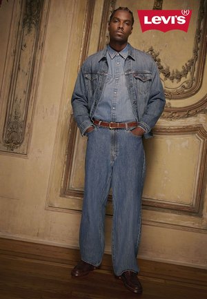 Lichtblauwe denimjas en bijpassend hemd met wijde jeans, bruine riem en donkere schoenen, tegen een sierlijke vintage achtergrond.