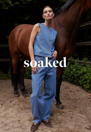 Donna con top di jeans senza maniche e jeans, in piedi accanto a un cavallo marrone in un'area sabbiosa all'aperto con vegetazione e recinzione in legno.