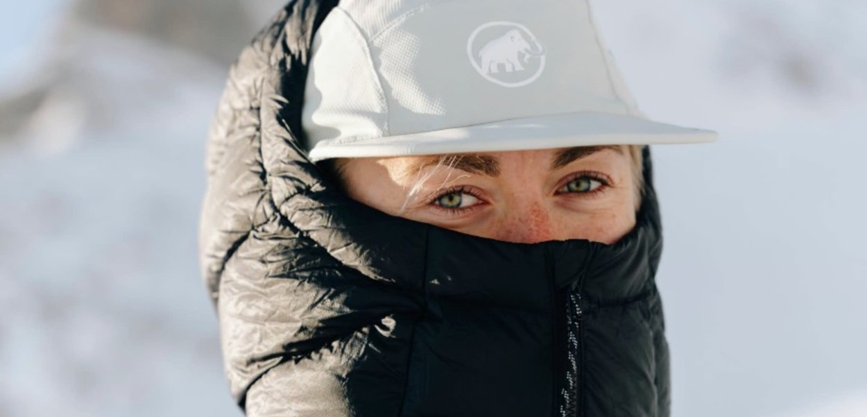 Schwarze Daunenjacke mit gestepptem Muster, hoher Kragen. Hellgraue Mütze mit Logo. Grüne Augen sichtbar, heller Hautton mit Sommersprossen.
