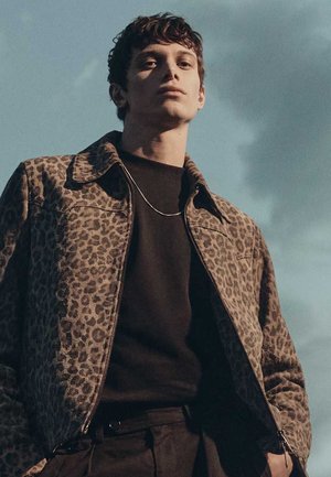 Młody mężczyzna w kurtce w panterkę i czarnej koszuli, stojący na zewnątrz na tle pochmurnego nieba.