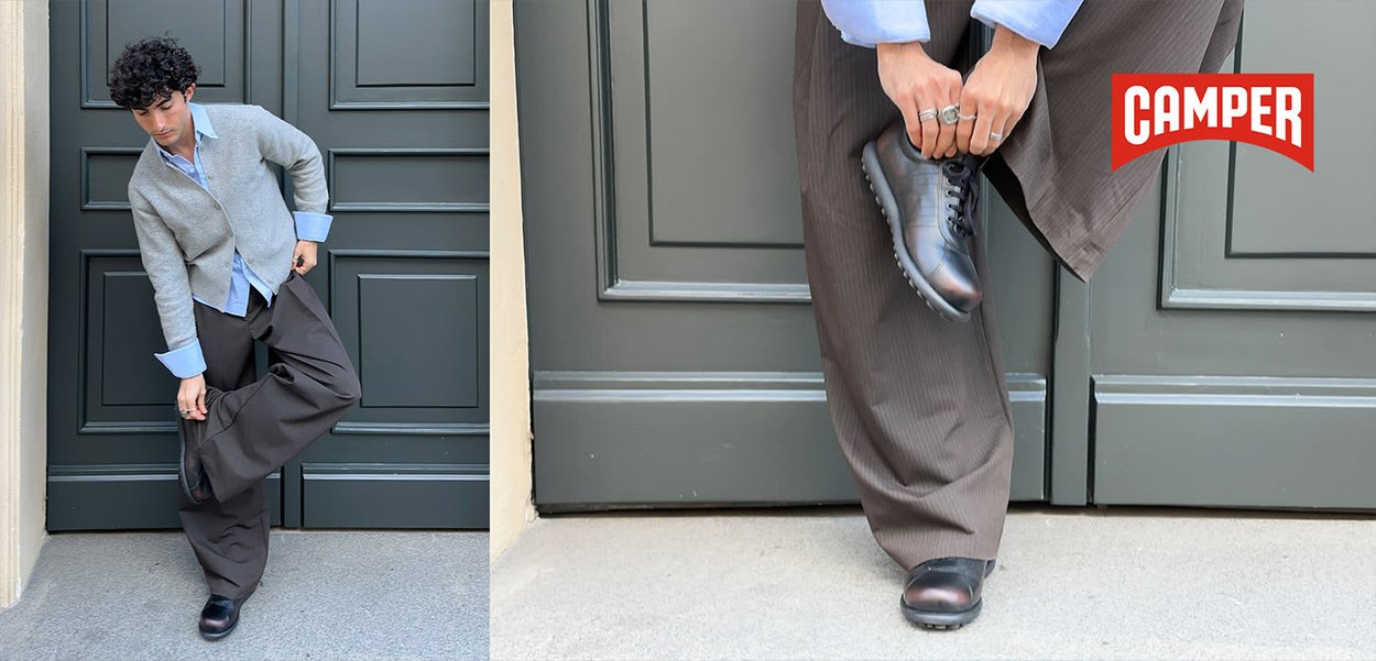 Man wearing gray cardigan and brown trousers puts on black leather shoes in front of gray paneled door, Camper logo visible.