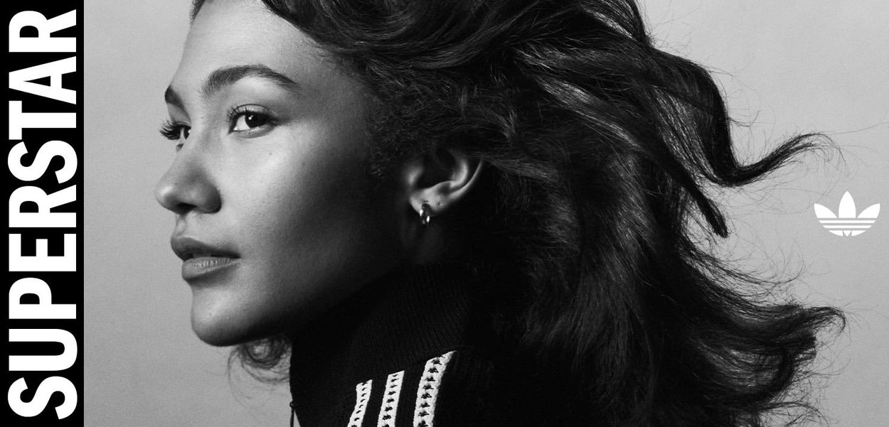 Mujer con cabello suelto y chaqueta Adidas negra mirando hacia la izquierda, texto "SUPERSTAR" en vertical a la izquierda, logotipo de trébol de Adidas a la derecha.