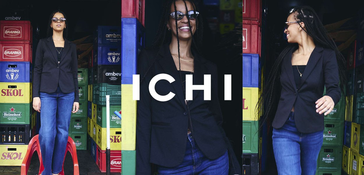 Frau mit langen Zöpfen und Sonnenbrille trägt schwarzen Blazer und Jeans, steht lächelnd zwischen gestapelten bunten Getränkekisten.