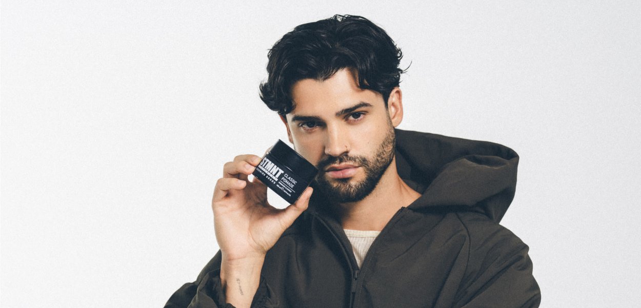 Zwarte pot met haarpomade wordt vastgehouden door een persoon die een donkerbruine jas met kap draagt. De pot heeft witte tekst op een zwarte achtergrond.