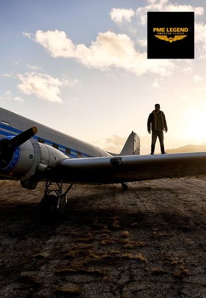 Metalen zilverkleurig vintage vliegtuig met blauwe strepen; een persoon staat op de vleugel. Zonsondergang op de achtergrond; grasbedekte grond. PME Legend-logo.
