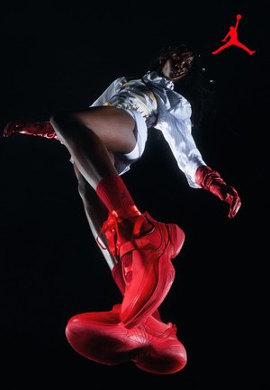 Osoba nosząca czerwone trampki, czerwone rękawiczki, czerwone skarpetki, białe szorty i białą koszulę w wzory, skacząca na czarnym tle z czerwonym logo.