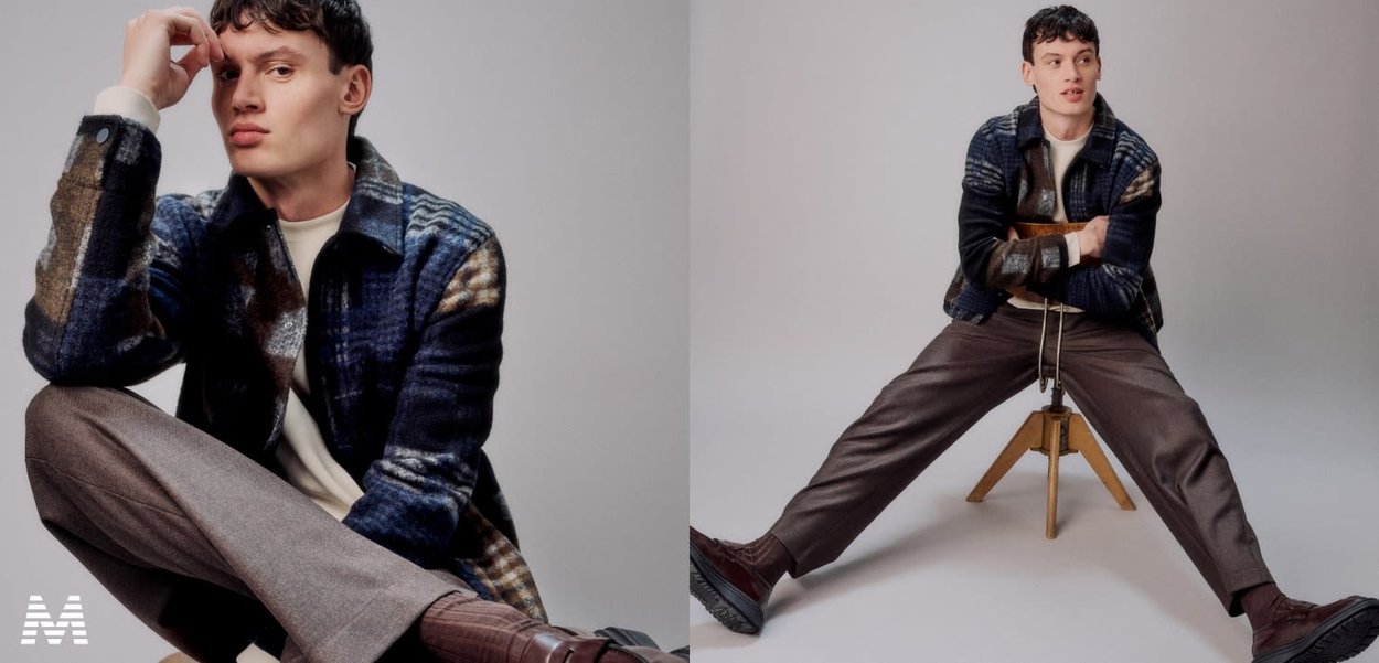 Menswear: blue and brown patterned wool jacket over a white shirt, grey trousers, and brown shoes. Model seated with relaxed pose.