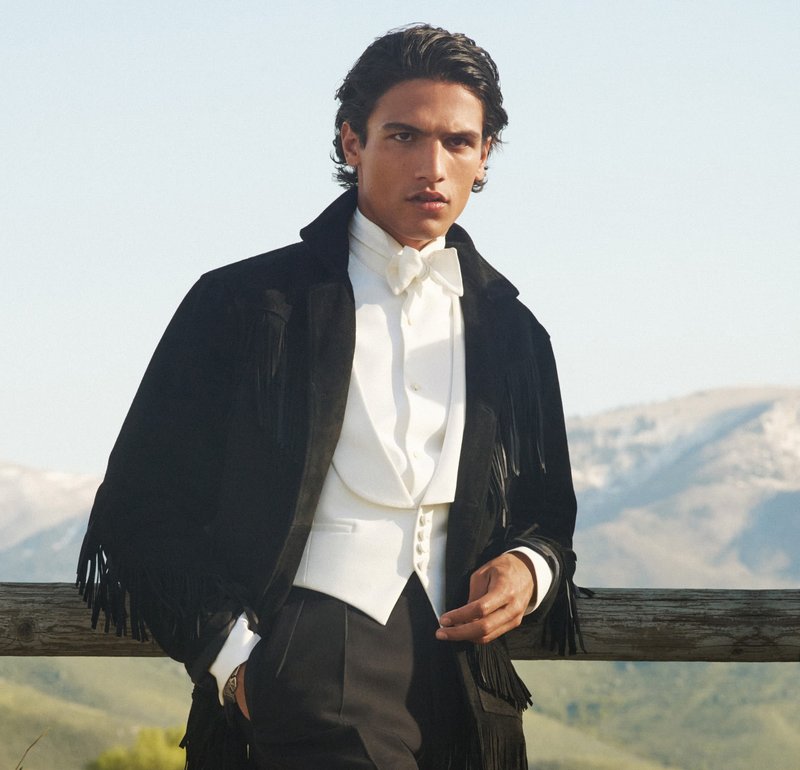 Veste en daim noire avec des détails en franges, gilet blanc et nœud papillon. Le modèle se tient devant un fond montagneux.