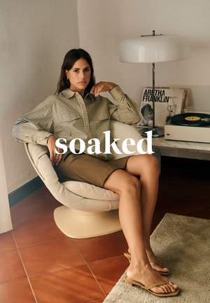 Kariertes Hemd mit zwei Fronttaschen, beige Rock und hellbraune Sandalen. Gebogener Stuhl, Plattenspieler und weicher Teppich in hellem Raum.