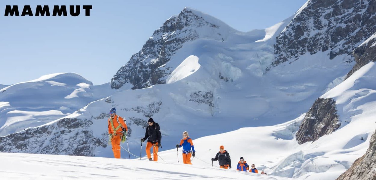 Sechs Bergsteiger sind mit Seilen verbunden und wandern über schneebedecktes Gelände, während im Hintergrund zerklüftete, schneebedeckte Berge zu sehen sind.